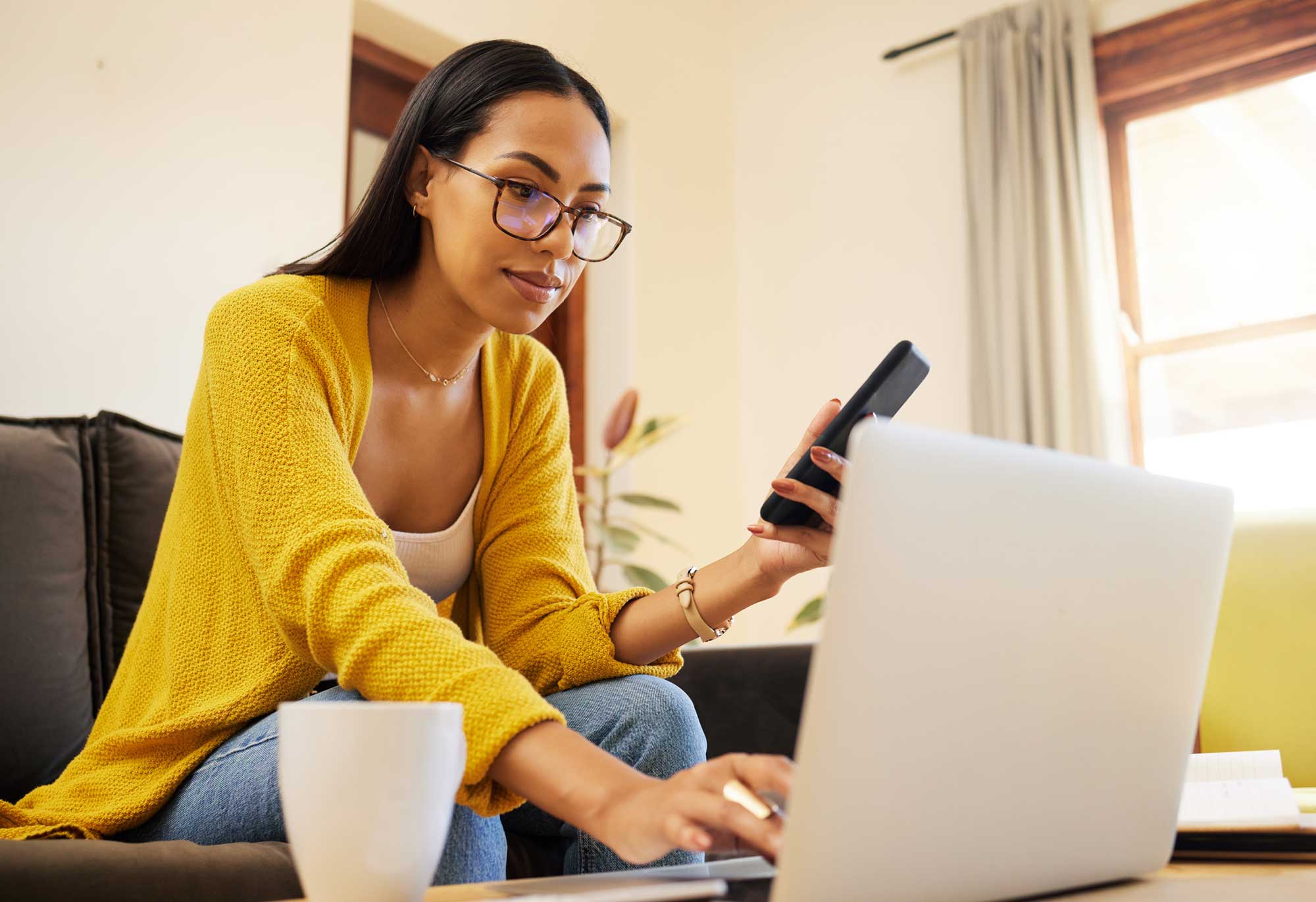young women with glasses working on a laptop and holding a mobile phone.